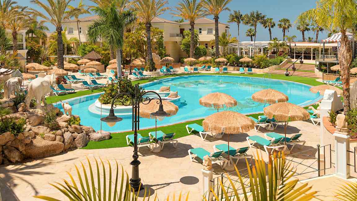 Kuva ylellisestä lomakohteesta, jossa on uima-allas aurinkotuolien ja aurinkovarjojen ympäröimänä, palmuja ja rakennuksia ympärillä. (Tekoälyn luoma teksti)