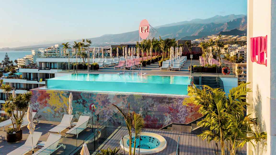 Suuri kattoterassi, jossa on uima-allas, aurinkotuoleja ja kasveja. Taustalla vuoria ja rakennuksia. (Tekoälyn luoma teksti)