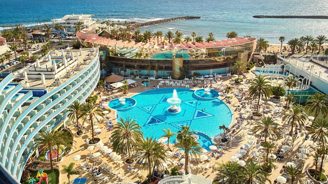 Näkymä lomakeskuksesta, jossa on allasalue, aurinkotuoleja ja palmuja lähellä merta. (Tekoälyn luoma teksti)