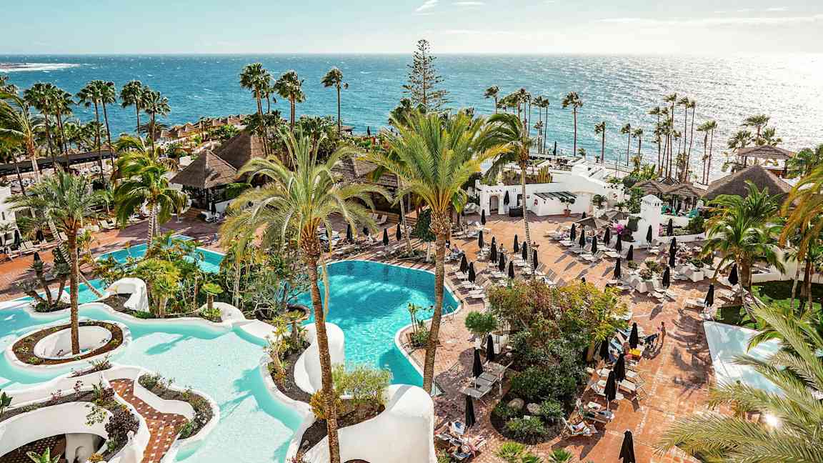 Näköala hotellialueelle, jossa on kaksi uima-allasta, palmuja ja aurinkotuoleja meren rannalla. (Tekoälyn luoma teksti)