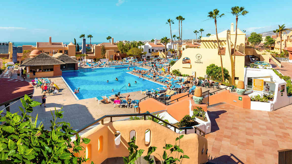 Hotellialue, jossa on suuri uima-allas, aurinkotuoleja ja rakennuksia. (Tekoälyn luoma teksti)