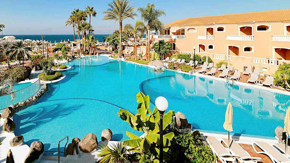 Suuri uima-allas, jota ympäröivät aurinkovarjot, aurinkotuolit, palmut ja taustalla oleva rakennus. (Tekoälyn luoma teksti)