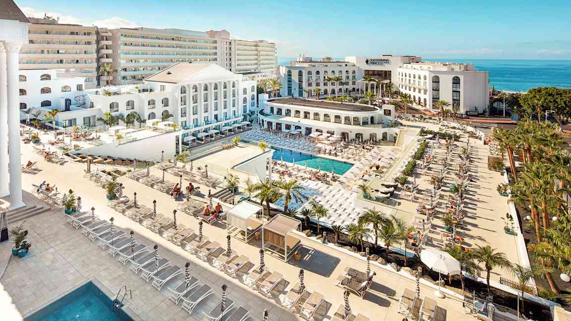 Näkymä hotellialueelle, jossa on uima-altaita, aurinkotuoleja ja vaalea arkkitehtuuri meren lähellä. (Tekoälyn luoma teksti)