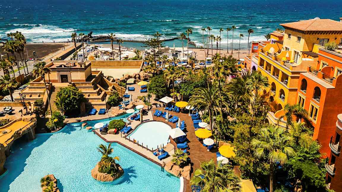 Hotellialue, jossa on uima-altaita, kasvillisuutta ja värikkäitä rakennuksia meren rannalla. (Tekoälyn luoma teksti)