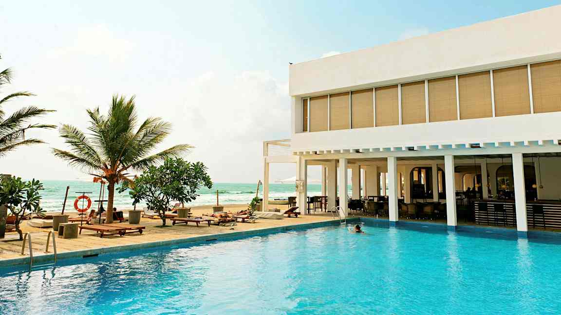 Ulkouima-allas, jonka vierellä on aurinkotuoleja ja palmuja modernin hotellin vieressä meren äärellä. (Tekoälyn luoma teksti)