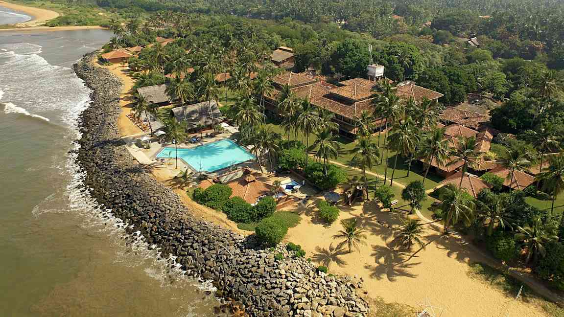 Näkymä rannikkoviivalle, jossa hotellialue, johon kuuluu uima-allas, tiilikattoisia rakennuksia sekä vehreitä ja kookospuiden ympäröimiä maisemia. (Tekoälyn luoma teksti)