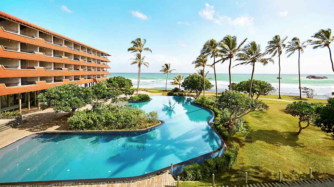 Hotellirakennus rannalla, jossa on suuri uima-allas, palmuja ja meri taustalla. (Tekoälyn luoma teksti)