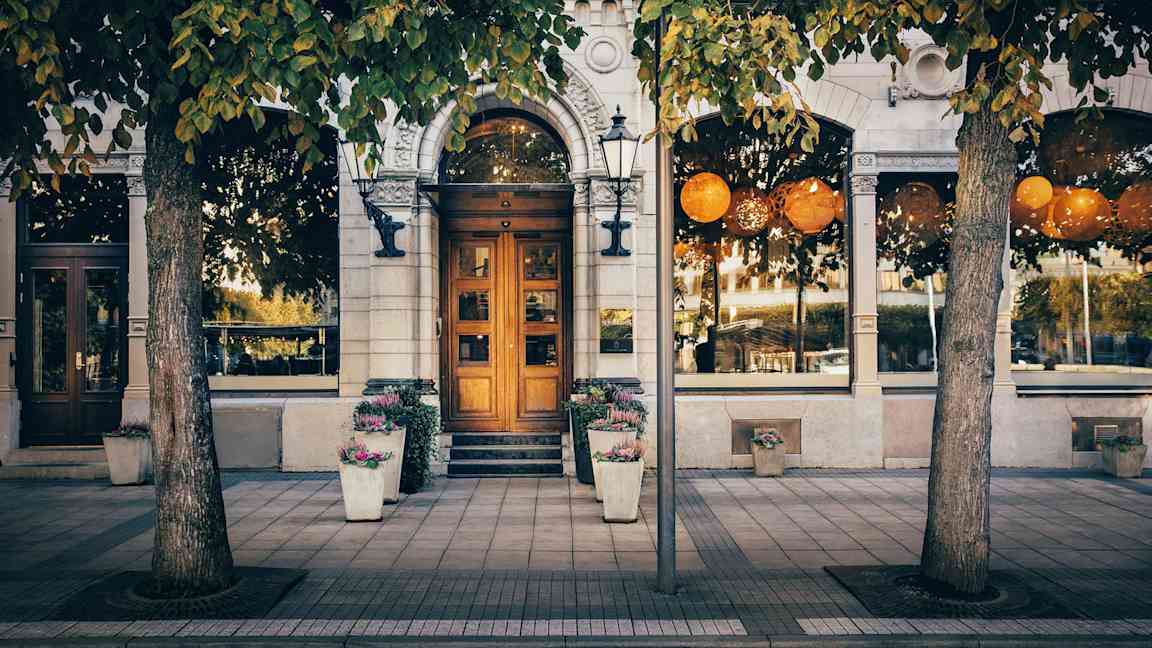 Rakennuksen julkisivu, jonka edessä on puita, suuria ikkunoita, iso puuovi ja kukkaruukkuja jalkakäytävällä. (Tekoälyn luoma teksti)