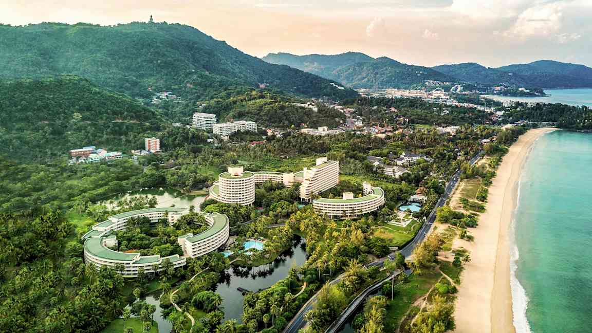 Ilmakuva rannikkoalueesta, jossa on hotellirakennuksia, ympäröivää vihreää ja ranta. (Tekoälyn luoma teksti)