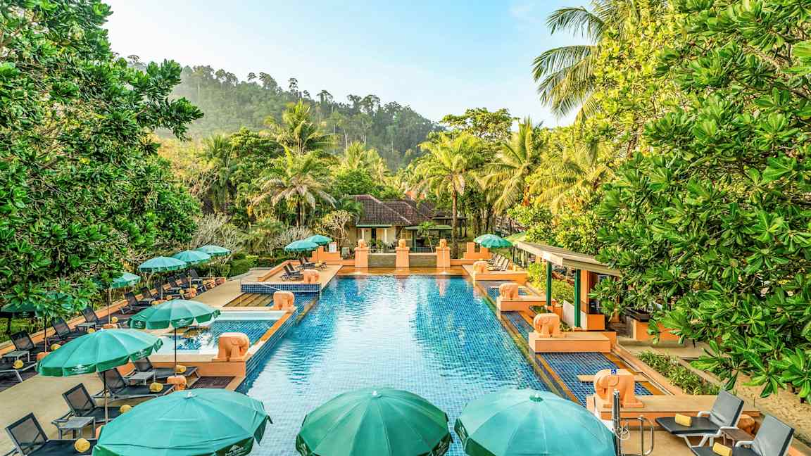 Ulkouima-allas, jossa sinistä vettä, ympäröitynä vihreillä aurinkovarjoilla ja aurinkotuoleilla, taustalla vihreä ympäristö ja huvila. (Tekoälyn luoma teksti)