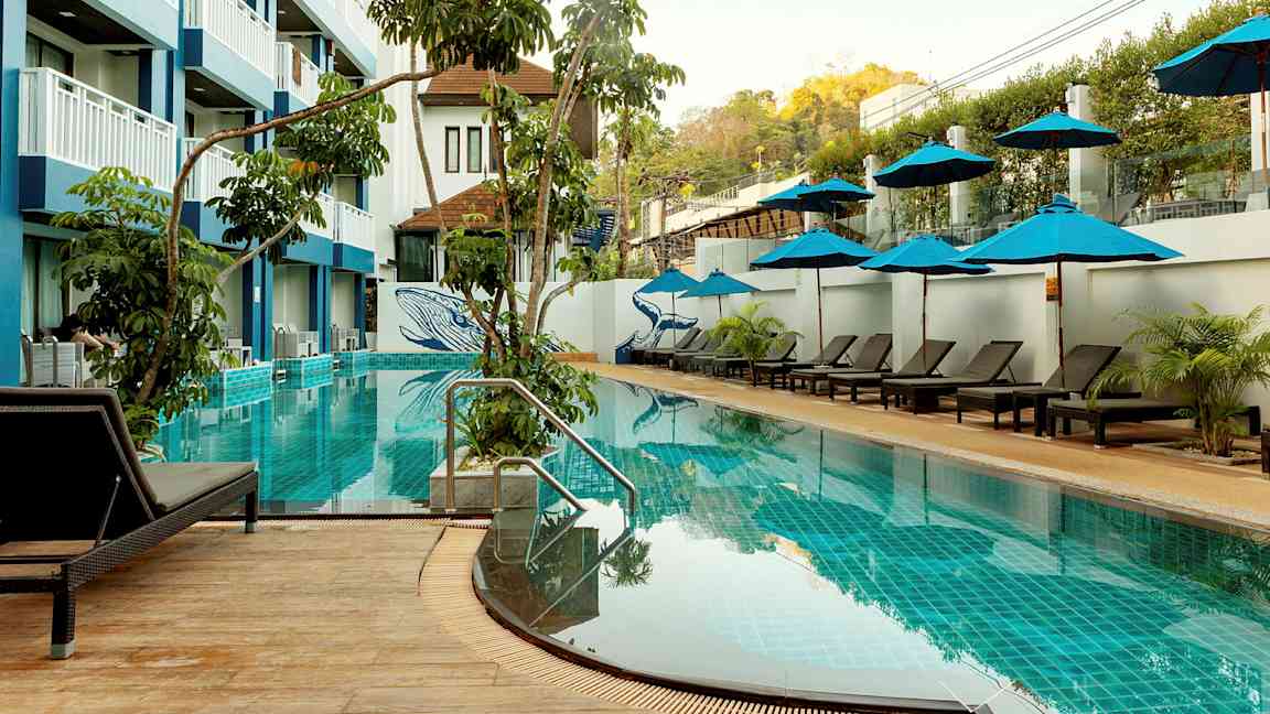 Ulkouima-allas, jossa aurinkotuoleja ja aurinkovarjoja, ympäröitynä kasvillisuudella ja monikerroksisella hotellirakennuksella. (Tekoälyn luoma teksti)