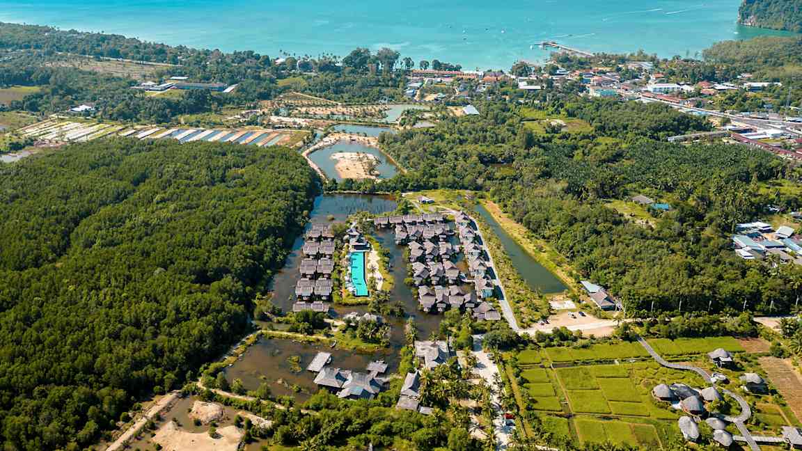 Ilmakuva lomakeskuksesta, jota ympäröi kasvillisuus; keskiössä uima-allas ja taustalla meri. (Tekoälyn luoma teksti)