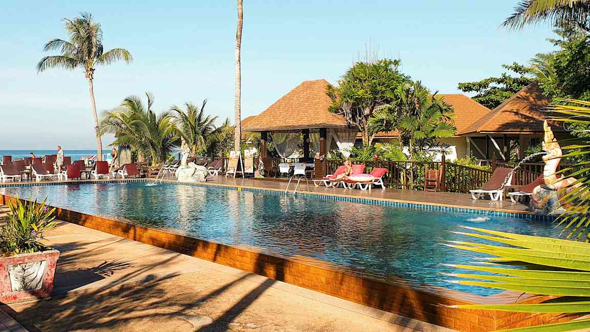 Kuva uima-allasalueesta, jossa on palmuja, aurinkotuoleja ja bungalow-tyylisiä rakennuksia taustalla. (Tekoälyn luoma teksti)