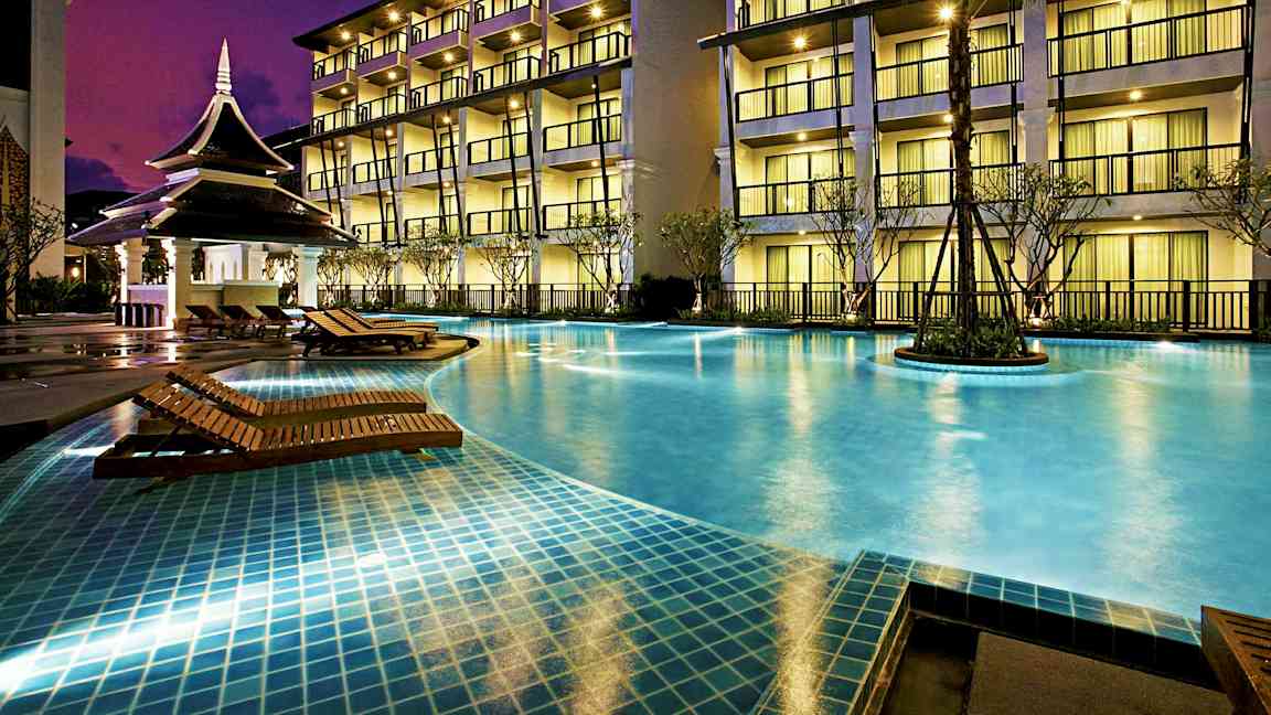 Valaistu hotellin julkisivu, jossa on parvekkeita useassa kerroksessa, edessä uima-allas puisine aurinkotuoleineen ja pieni paviljonki. (Tekoälyn luoma teksti)