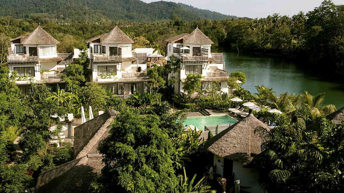 Hotellialue, jossa on trooppista tyyliä olevia taloja, joita ympäröi vehreys ja taustalla virtaava joki. (Tekoälyn luoma teksti)