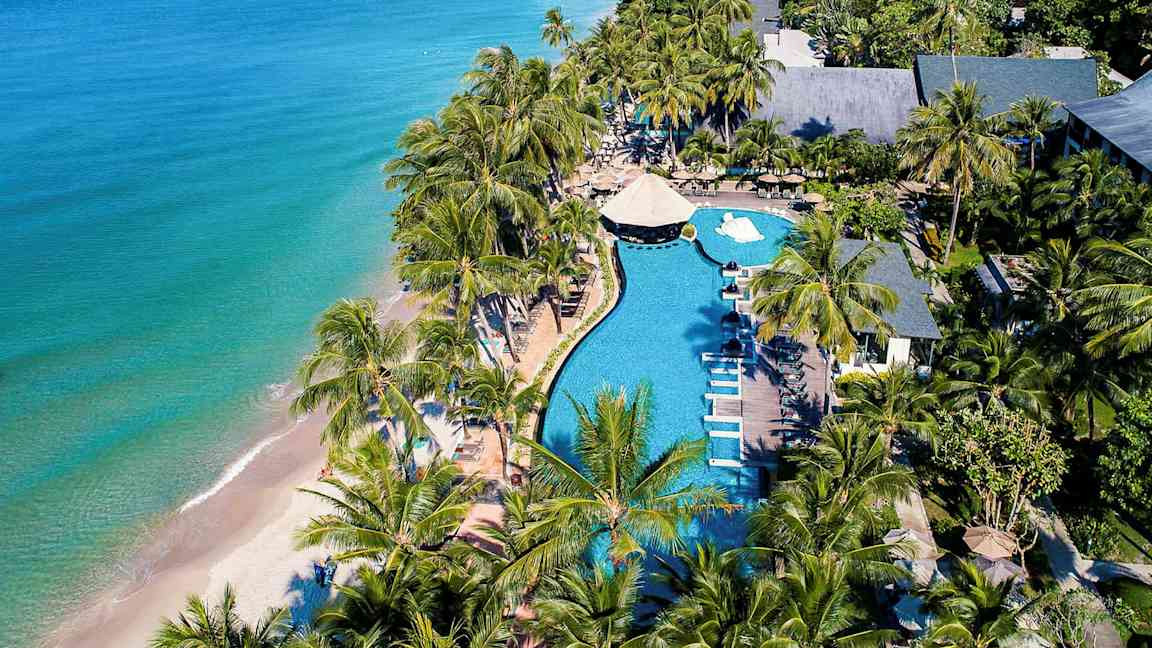 Ilmakuva rantakohteesta, jossa on uima-allas, aurinkotuoleja ja palmuja lähellä merta. (Tekoälyn luoma teksti)