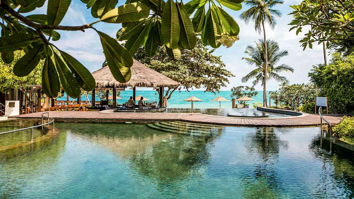 Suuri uima-allas trooppisessa ympäristössä, jossa on puita, palmuja ja meri taustalla. (Tekoälyn luoma teksti)