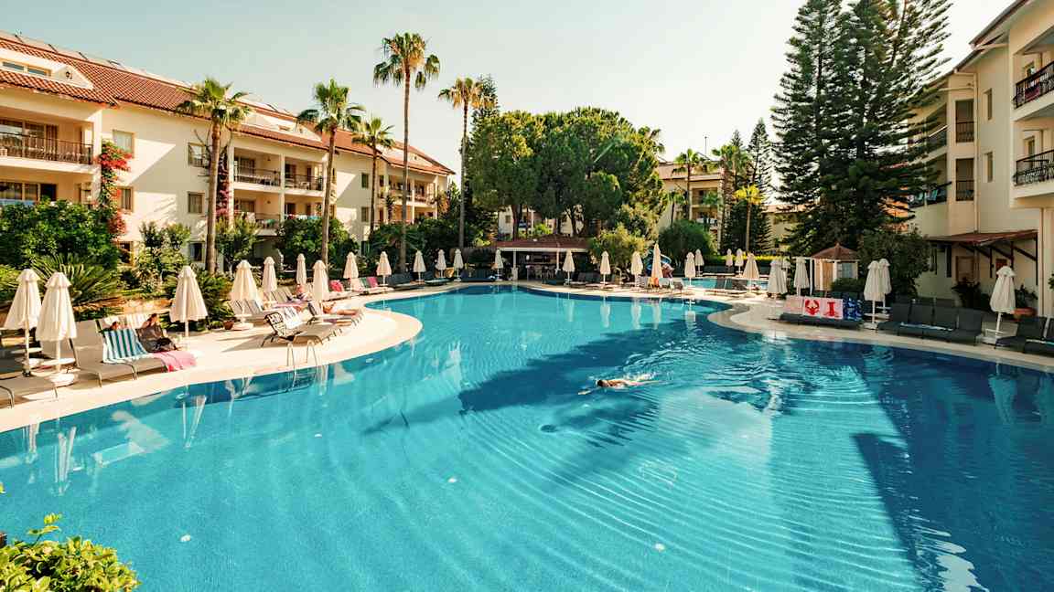 Suuri ulkouima-allas, jota ympäröivät aurinkotuolit ja aurinkovarjot, taustalla vihreitä puita ja hotellirakennuksia. (Tekoälyn luoma teksti)