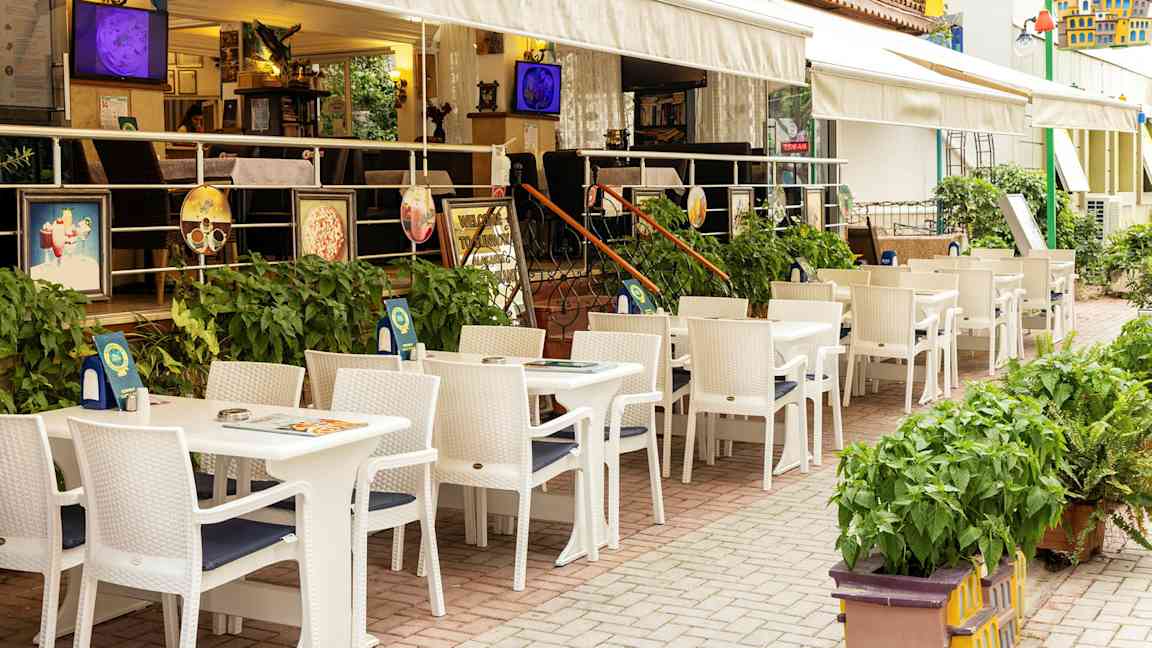 Ravintola ulkoterassilla, valkoiset pöydät ja tuolit hotellin vieressä. (Tekoälyn luoma teksti)