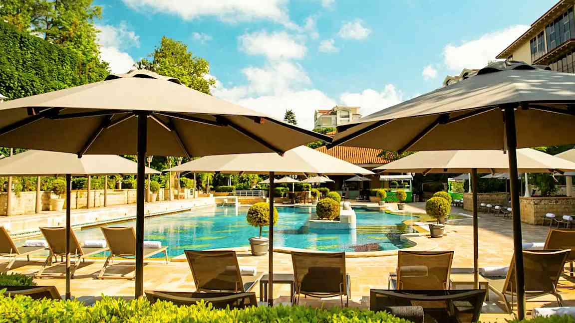 Uima-allasalue, jossa aurinkovarjoja, aurinkotuoleja ja ympäröivää vihreää. (Tekoälyn luoma teksti)