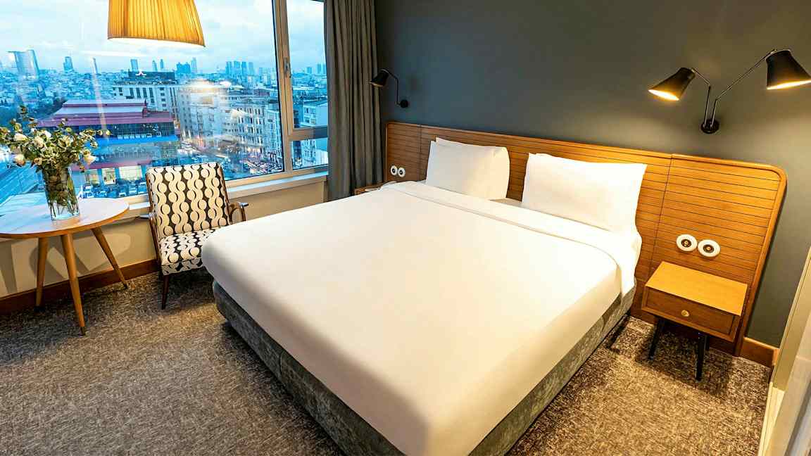 Hotellihuone, jossa suuri sänky, ikkuna ja näkymä kaupunkiin. (Tekoälyn luoma teksti)