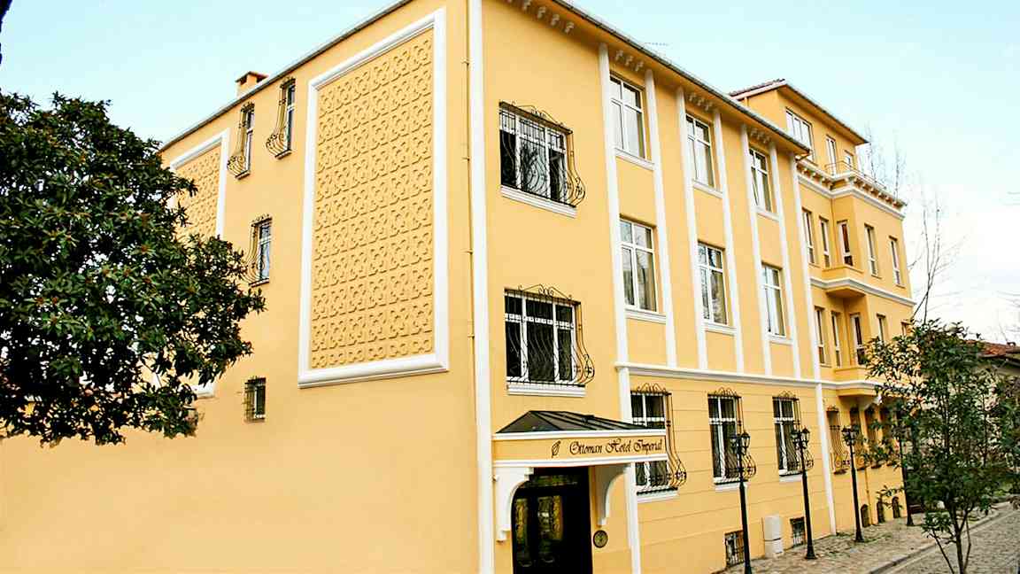 Keltainen hotelli, jossa on koristeellinen julkisivu, ikkunat rautaritilöillä ja sisäänkäynti katoksella. (Tekoälyn luoma teksti)