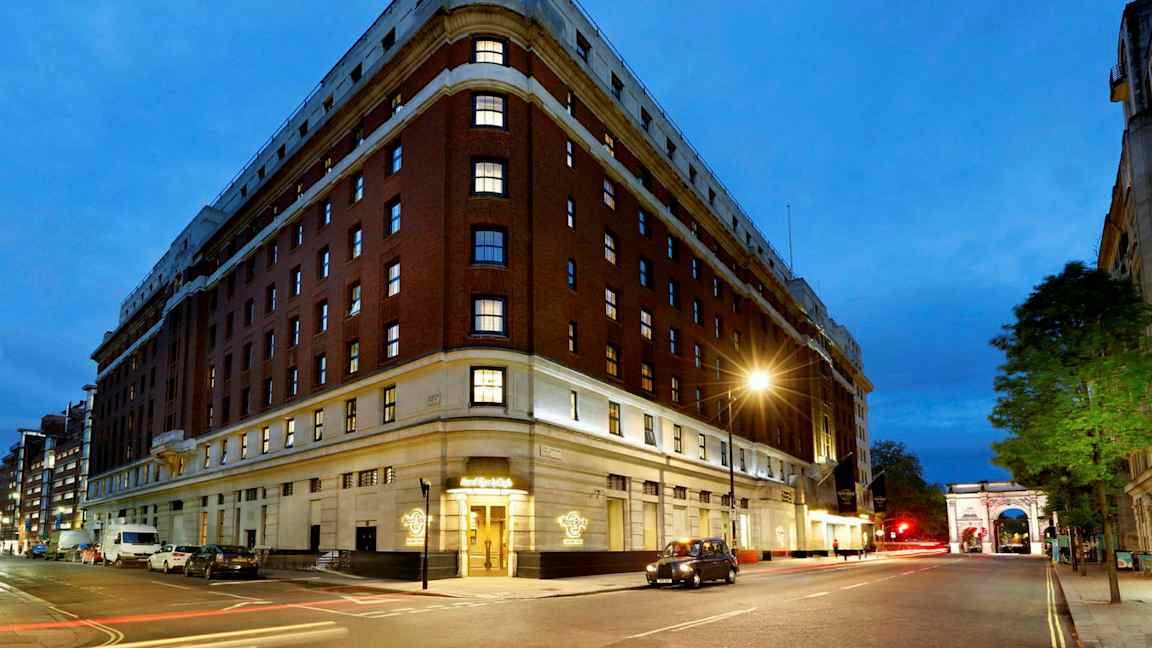 Suuri rakennus, jossa on punatiiliseinä ja useita kerroksia, näkyy yöllä katuvalojen ja liikennevalojen valossa. Kaari sijaitsee kadun varrella. (Tekoälyn luoma teksti)