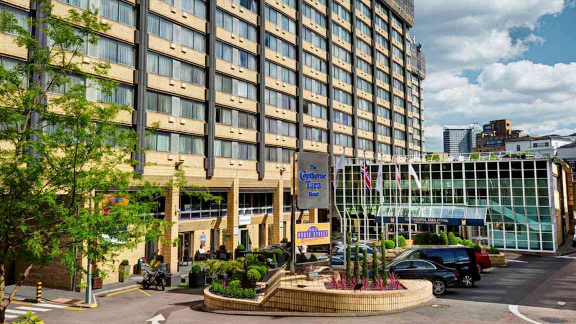 Monikerroksinen hotellirakennus, lasiovi ja pysäköintialue edessä. (Tekoälyn luoma teksti)