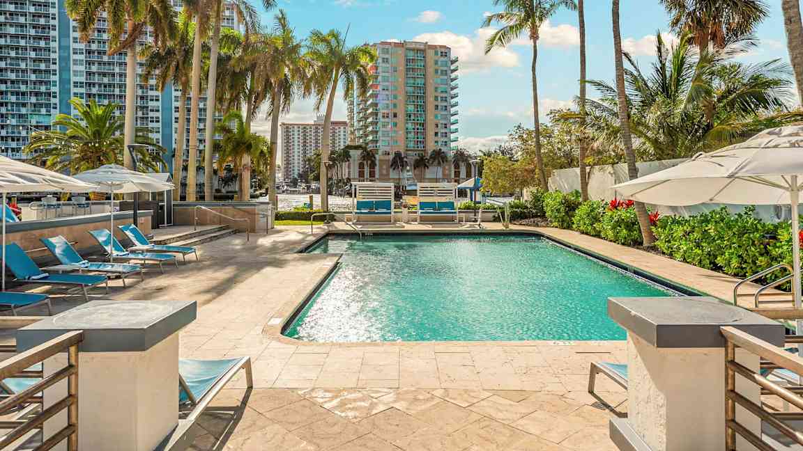 Uima-allas ympäröitynä aurinkotuoleilla ja aurinkovarjoilla, taustalla näkyy korkeita rakennuksia ja palmupuita. (Tekoälyn luoma teksti)