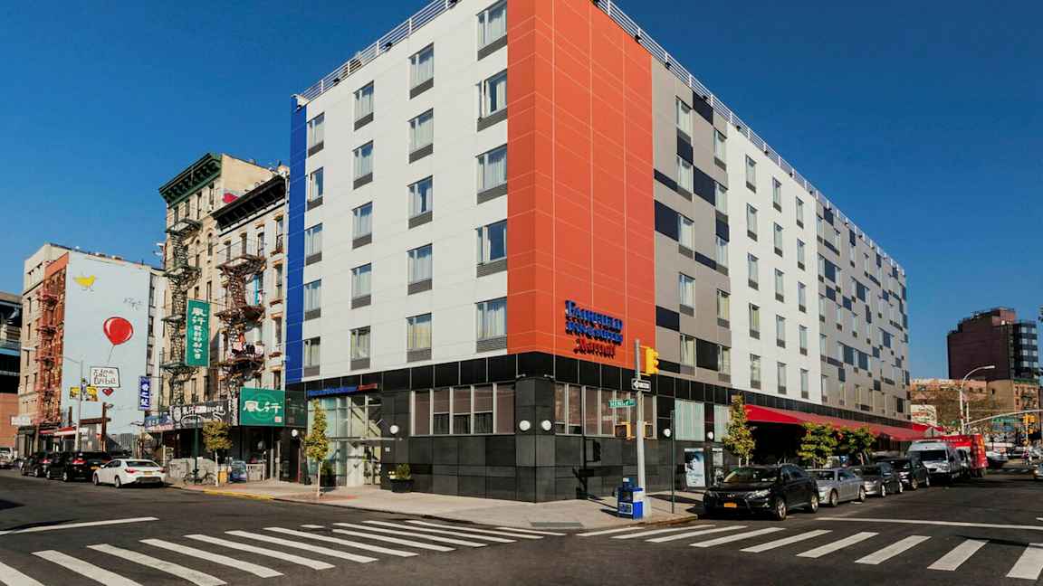 Moderni rakennus kaupunkialueen kulmassa, vilkas katu ja liikenne ympäröivien vanhempien rakennusten lomassa. (Tekoälyn luoma teksti)