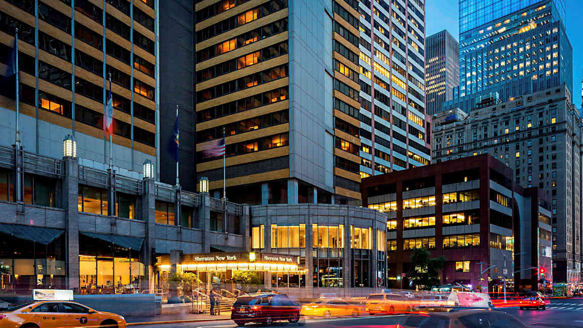 Kuva kaupunkimaisemasta, jossa on korkeita rakennuksia. Yhdessä rakennuksessa lukee "Sheraton New York" sen sisäänkäynnillä. Takseja näkyy kadulla. (Tekoälyn luoma teksti)