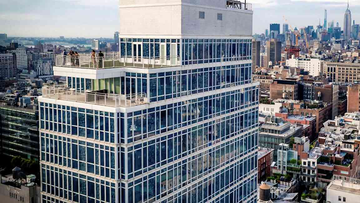 Korkea rakennus lasijulkisivulla ja terassilla, kaupunkimaiseman ympäröimänä. (Tekoälyn luoma teksti)