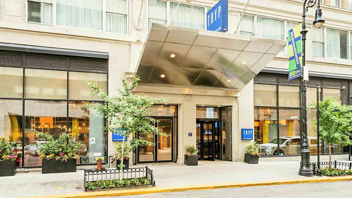 Hotellin sisäänkäynti, jossa on kyltti "TRYP". Rakennuksessa on suuret ikkunat ja istutuksia. (Tekoälyn luoma teksti)