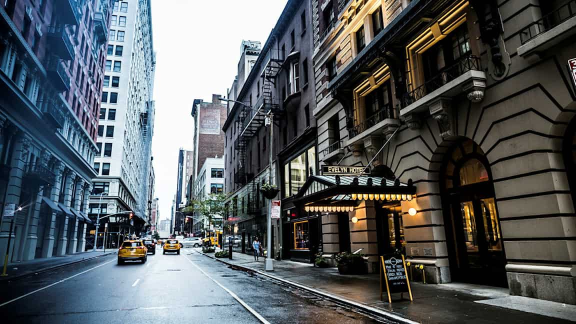 Kaupunkikatu, jonka molemmin puolin on korkeita rakennuksia, ja tiellä liikkuu takseja. Evelyn-hotellin kyltti näkyy oikealla. (Tekoälyn luoma teksti)