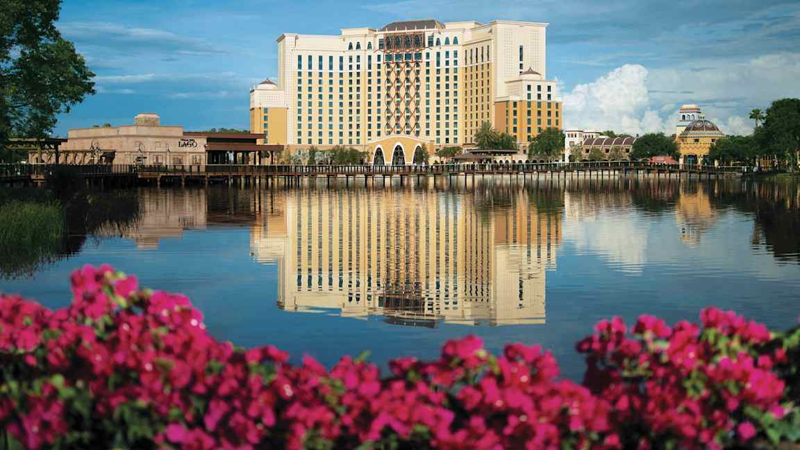 Hotellikompleksi heijastuu rauhalliseen järveen, etualalla on vaaleanpunaisia kukkia. (Tekoälyn luoma teksti)