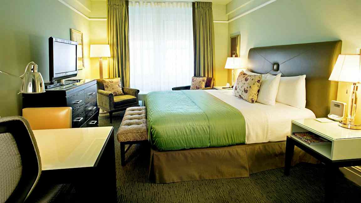 Hotellihuone, jossa on suuri sänky, työpöytä ja televisio. Huone on sisustettu vihrein sävyin ja siinä on suuret ikkunat. (Tekoälyn luoma teksti)