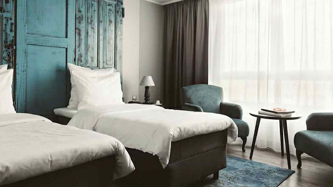 Hotellihuone, jossa on kaksi erillistä vuodetta, sinisiä yksityiskohtia seinässä, pöytä ja nojatuoleja sekä suuri ikkuna valkoisella verholla. (Tekoälyn luoma teksti)