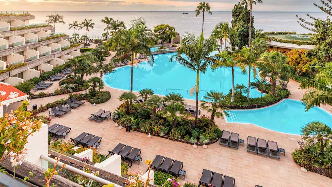 Kuva hotellikompleksista, jossa on suuri uima-allas, ympäröity aurinkotuoleilla ja trooppisilla kasveilla. Taustalla näkyy meri ja auringonlasku. (Tekoälyn luoma teksti)