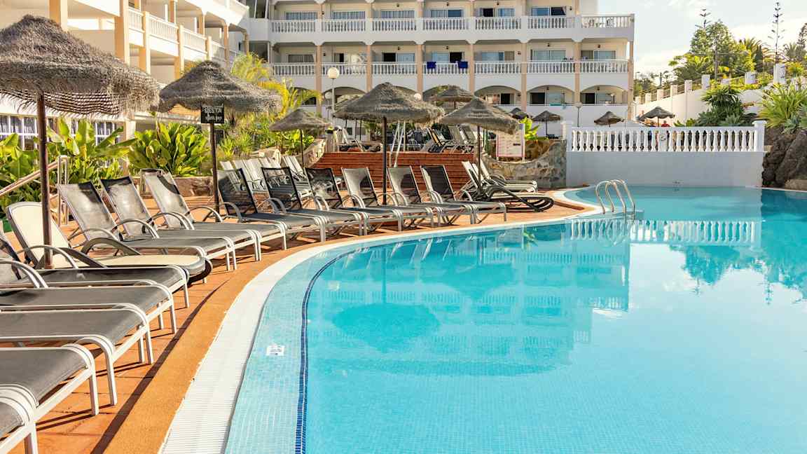 Kuvassa monikerroksinen hotellirakennus uima-altaan vieressä. Altaan edessä lepotuoleja ja aurinkovarjoja. (Tekoälyn luoma teksti)