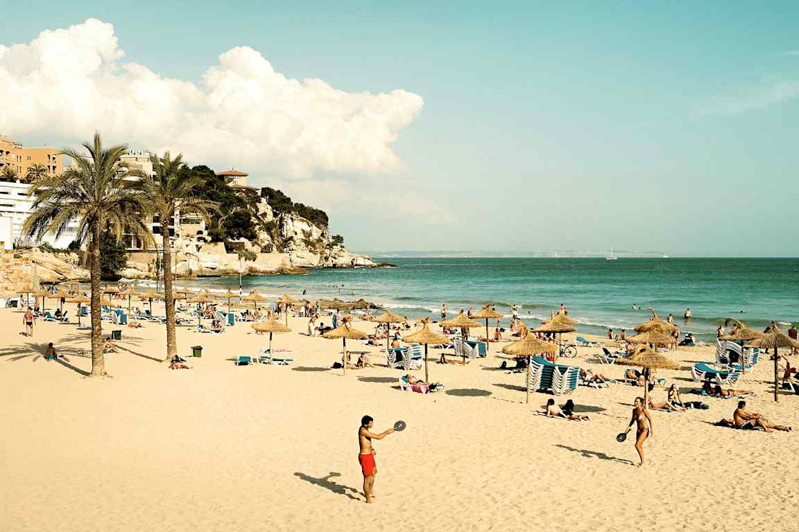 Hiekkaranta, jossa on aurinkovarjoja, aurinkotuoleja ja ihmisiä nauttimassa auringosta ja merestä. Kauempana näkyy kallio ja rakennuksia. (Tekoälyn luoma teksti)