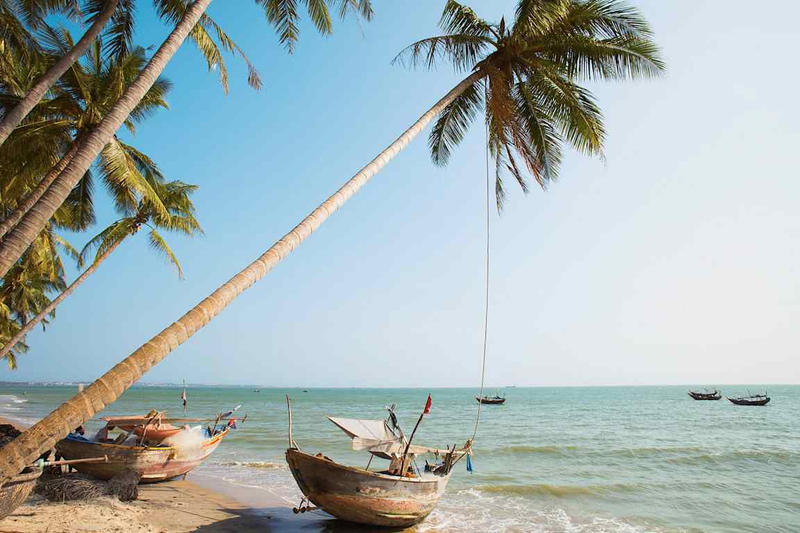 Näkymä trooppiselle rannalle, jossa palmuja ja perinteisiä veneitä veden äärellä. (Tekoälyn luoma teksti)