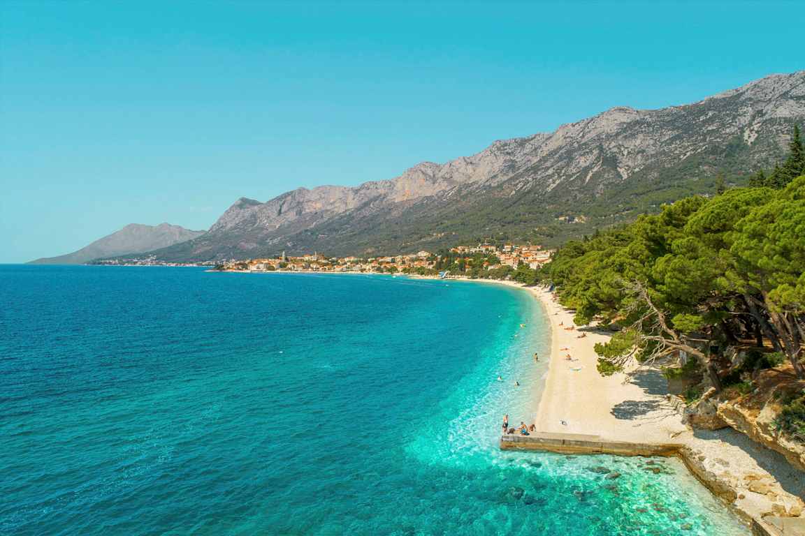 Näkymä kivirannalle, jossa on turkoosia vettä ja vehreitä puita. Taustalla näkyy vuoria ja kylä. (Tekoälyn luoma teksti)