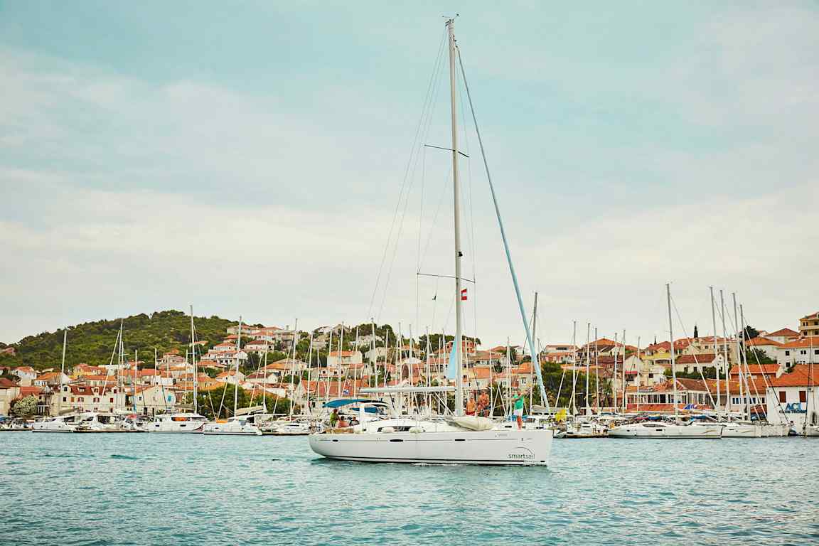 Valkoinen purjevene satamassa, taustalla rakennuksia ja vihreitä kukkuloita. (Tekoälyn luoma teksti)