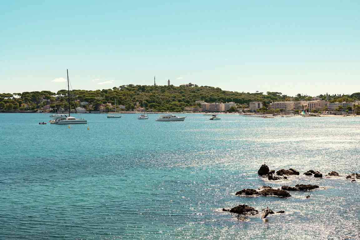 Näkymä rantaviivasta, jossa on kirkasta sinistä vettä, useita veneitä ja purjeveneitä vehreän rannan ja kaupungin läheisyydessä. (Tekoälyn luoma teksti)