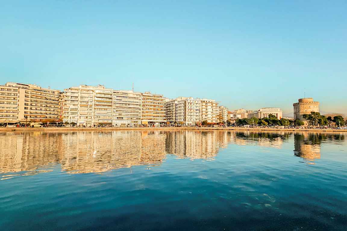 Kaupunki veden äärellä, jossa rakennuksia ja pyöreä torni heijastuvat tyynessä vedessä. (Tekoälyn luoma teksti)