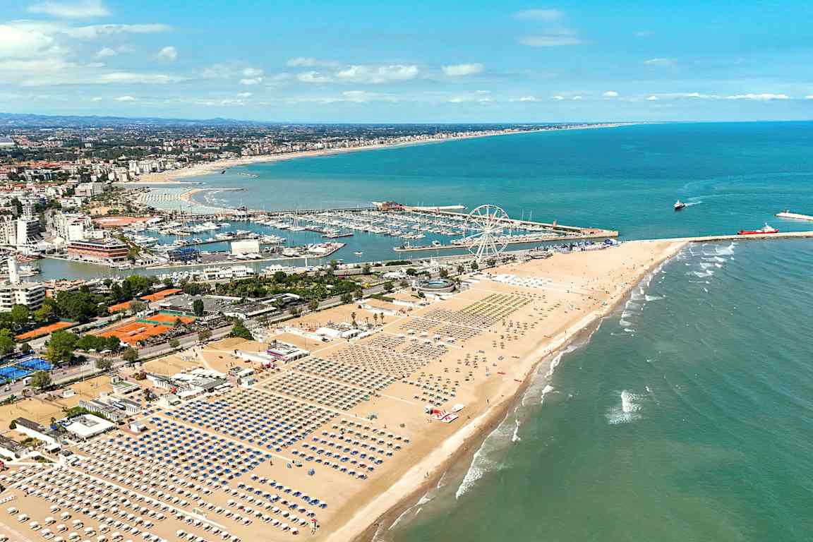 Näköala leveälle hiekkarannalle, jossa on rivi toisensa jälkeen aurinkotuoleja ja sateenvarjoja. Taustalla näkyy venesatama ja huvipuistomainen alue, jolla on suuri maailmanpyörä. Meri avautuu oikealla puolella. (Tekoälyn luoma teksti)