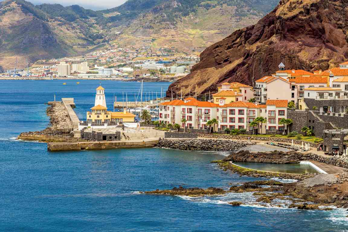 Rannikko, jossa punakattoisia rakennuksia, satama ja taustalla mäkiä. (Tekoälyn luoma teksti)