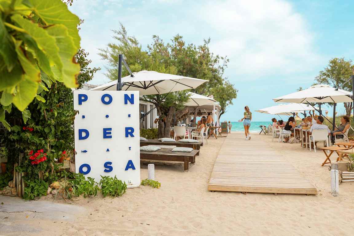 Ravintola rannalla, jossa on pöytiä ja aurinkovarjoja. Puiset kulkuväylät johtavat kohti vettä. (Tekoälyn luoma teksti)