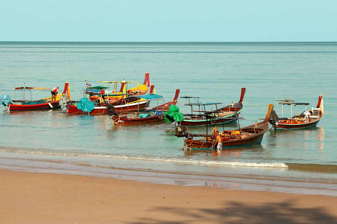 Useita värikkäitä veneitä on kiinnitettynä veteen lähellä hiekkarantaa. (Tekoälyn luoma teksti)