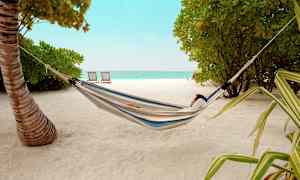 Henkilö lepää riippumatossa hiekkarannalla, taustalla puita ja meri. (Tekoälyn luoma teksti)